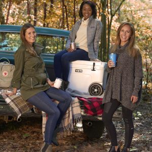 Three women tailgate by a truck in the woods.